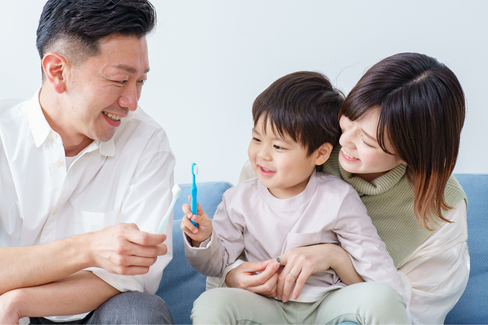 A smiling mother and father sitting on a couch with their young son, holding toothbrushes together.